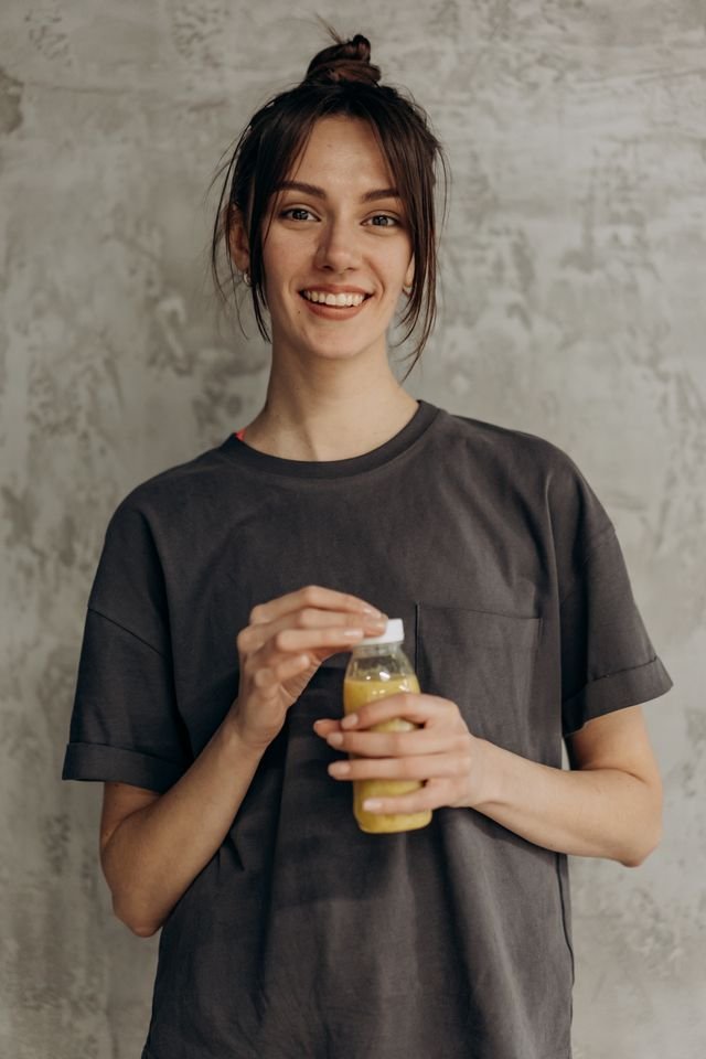 Casually dressed woman smiling and holding a juice, representing a healthy lifestyle indoors.